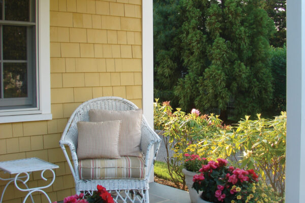 Colonial Yellow solid color prefinished cedar shingle siding on a covered porch wall with white trim, next to a window and patio seating.