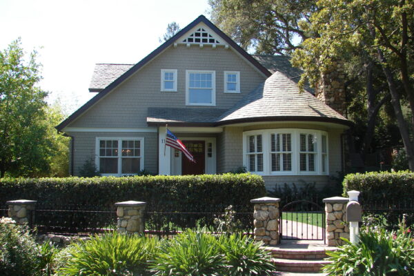 Dune Gray solid color prefinished cedar shingle siding on a cottage-style home with white trim, a curved bay window, and a dark shingle roof.