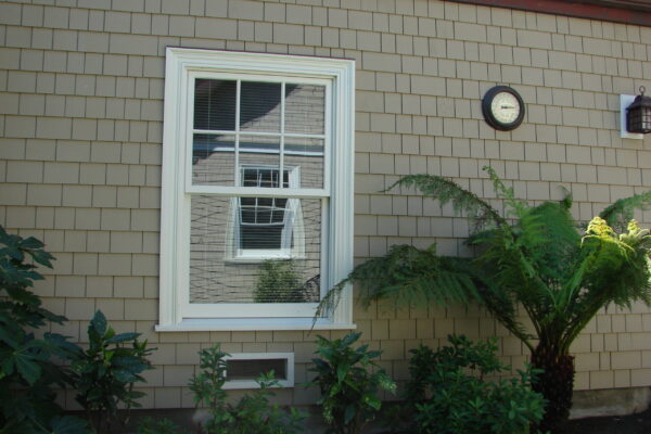 Dune Gray solid color prefinished cedar shingle siding on a cottage home with white trim, stone chimney, and a brick walkway through landscaped garden beds.