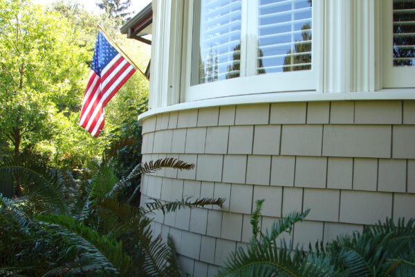 Dune Gray solid color prefinished cedar shingle siding on a curved bay window wall with white trim, with an American flag and landscaping in front.