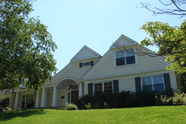 Linen solid color prefinished cedar shingle siding on a two-story home with white trim, black shutters, and a red front door, viewed from the front yard.