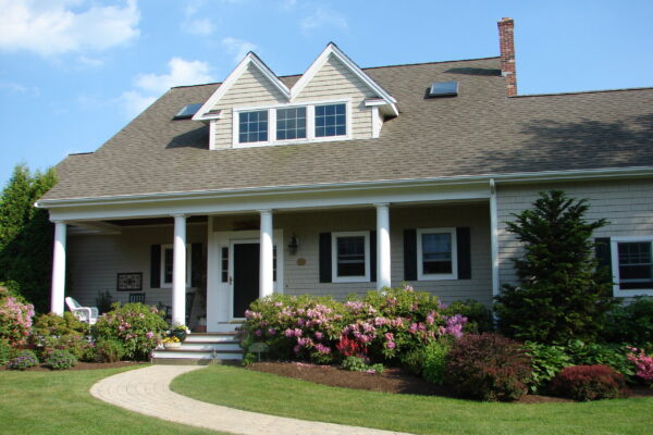 navajo-white-solid-color-prefinished-cedar-shingle-siding-white-columns-porch Navajo White solid color prefinished cedar shingle siding on a cottage-style home with white trim, a full-width porch with white columns, and a dark shingle roof.