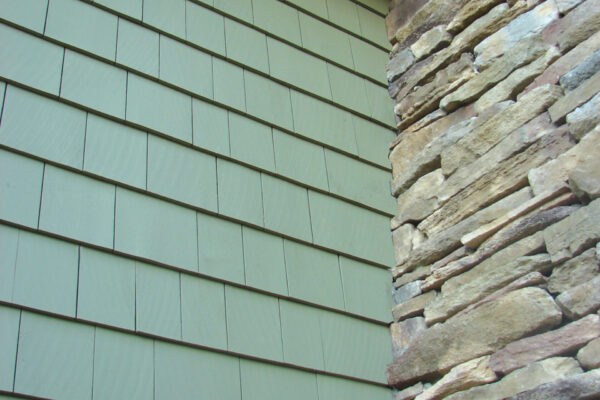 Sycamore solid color prefinished cedar shingle siding meeting a stacked-stone chimney, shown under a white soffit and trim.