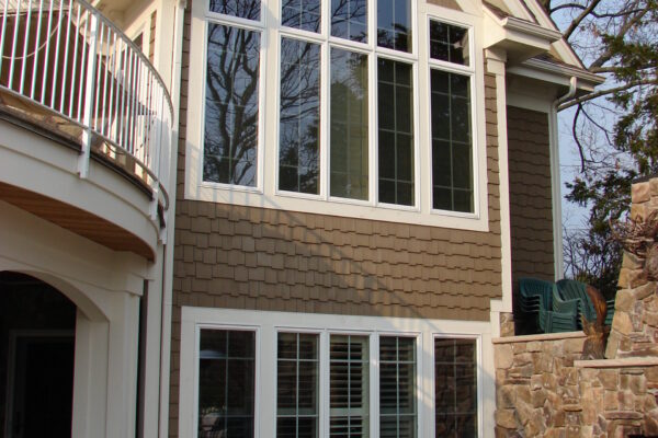 Taupe solid color prefinished cedar shingle siding on a gable-front home with white trim, a dark shingle roof, and a stone chimney.