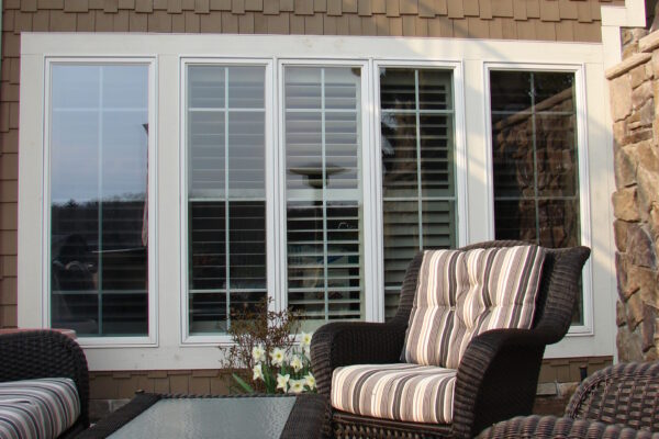 Taupe solid color prefinished cedar shingle siding above a row of large white-trim windows, with a stone wall edge beside a patio seating area.