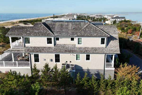 Aerial view of a coastal home clad in light gray cedar shingle siding with white trim and a weathered gray shingle roof, with ocean and dunes behind.