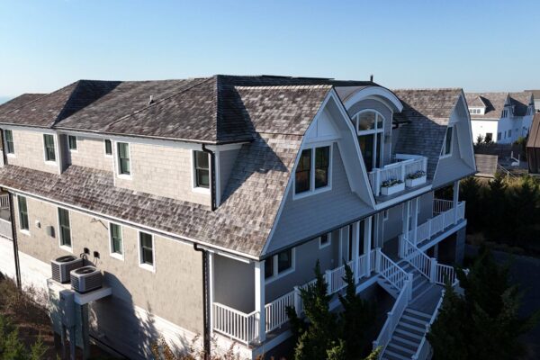 Aerial view of a coastal home with light gray cedar shingle siding, white trim, and a weathered gray shingle roof with a curved gambrel-style gable and balconies.