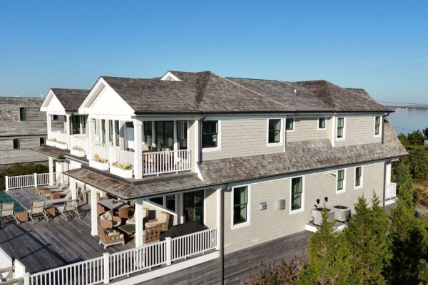 Aerial view of a coastal home with light gray cedar shingle siding, white trim, and wraparound decks, with water visible in the background.