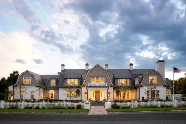 Wide front exterior of a large home with gray cedar shingle siding, white trim, and a dark shingle roof, photographed at dusk with warm interior lights.