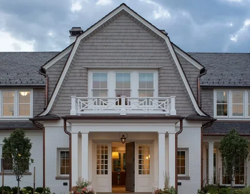Centered front entry of a home with gray cedar shingle siding, white trim, and a white-column porch with balcony above, photographed at dusk.