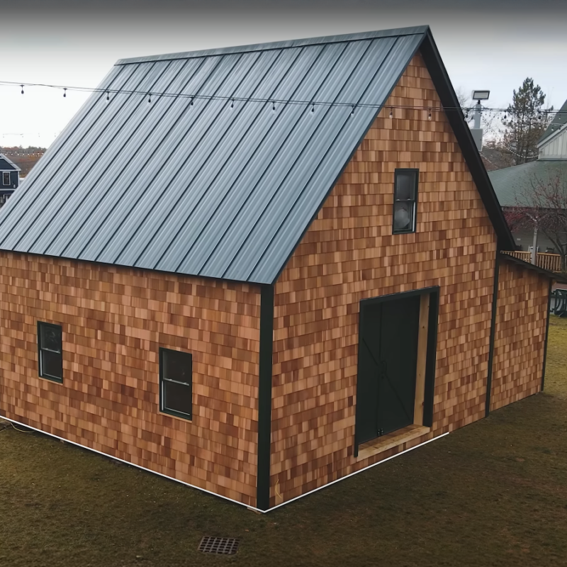 Small barn-style building clad in natural cedar shingle siding with a dark standing-seam metal roof, shown from an angled exterior view on a grassy lot.