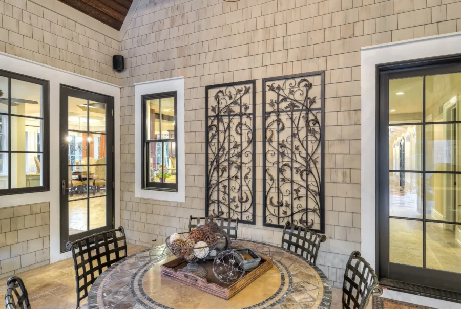Covered patio with light cedar shingle siding, black-trim glass doors and windows, and decorative wrought-iron wall panels above a round tile table.