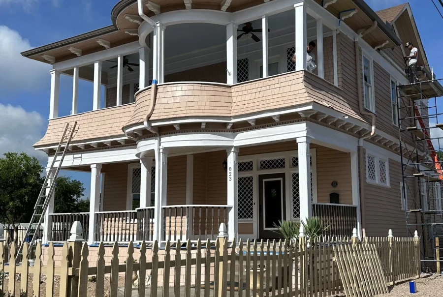 Two-story home with light tan cedar shingle siding, white trim, and wraparound porches; workers on scaffolding and a ladder indicate active exterior work.
