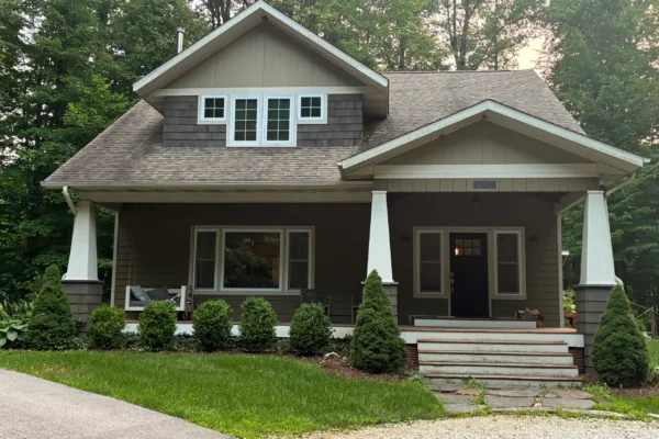 Front view of a craftsman-style home with dark gray cedar shingle siding, white tapered porch columns, tan gable accents, and a dark front door.