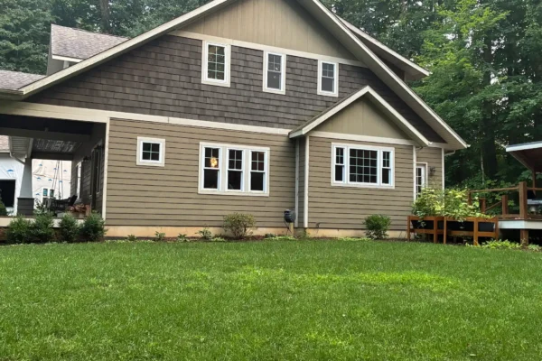 Rear view of a gable-roof house with dark gray cedar shingle siding on the upper story above tan lap siding, finished with white trim and multiple windows.