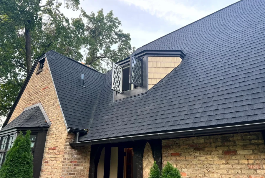 Dormer with light cedar shingle siding and a black-trim window set into a dark asphalt shingle roof above a brick exterior wall.