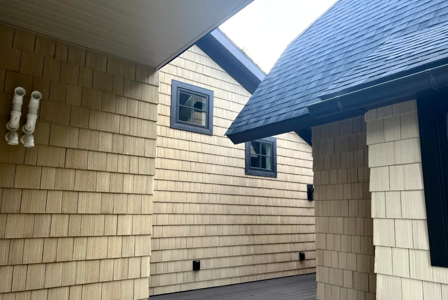Courtyard view of light cedar shingle siding with black-trim windows and a dark gray shingle roof overhang above a wood deck.