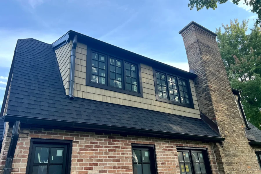Dormer clad in light natural cedar shingle siding with black-trim windows above a brick wall, with a dark gray shingle roof and a tall brick chimney.
