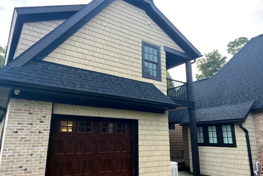 Garage and upper gable clad in light tan cedar shingle siding with black trim and a dark shingle roof, with a covered balcony and brick wall at left.
