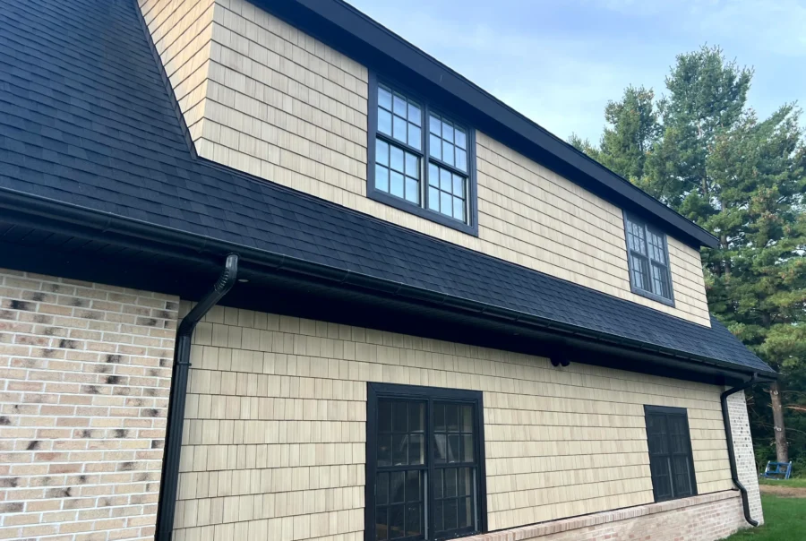Light tan cedar shingle siding with black-trim windows and black gutters on a long gable wall beneath a dark asphalt shingle roof.