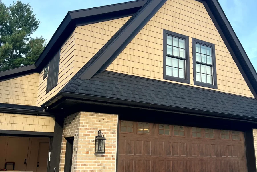 Light tan cedar shingle siding with black-trim windows and black gutters on a long home exterior wall above a light brick base.