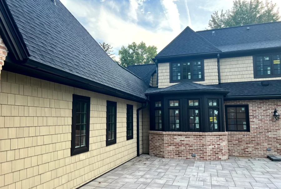 Courtyard view of a home with light tan cedar shingle siding, black-trim windows, black gutters, and a dark gray shingle roof above a brick section and paver patio.